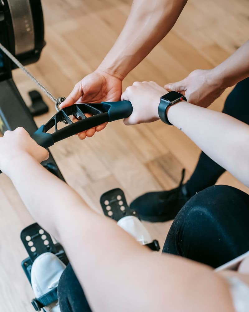 A coach's hands guide a client's grip on a rowing machine handle.